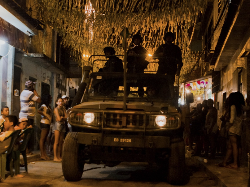 Still de filme. À noite, um veículo militar com os faróis acesos ocupa o centro de uma rua estreita, com homens fardados na parte superior. Moradores observam a movimentação nas calçadas e diante das casas. Acima da rua, numerosas fitas ou bandeirolas de festa decoram o espaço, iluminado por luzes em tons amarelados.