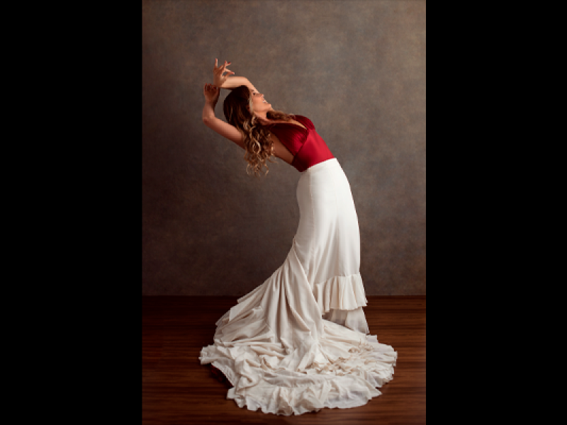 Retrato da bailarina Eliane Carvalho em pose de dança. Ela inclina o corpo para trás, com o rosto voltado para cima e os braços elevados e curvados sobre a cabeça. Veste blusa vermelha e uma longa saia branca com babados, cuja cauda se espalha pelo chão de madeira ao seu redor. Ao fundo, uma parede em tom marrom neutro destaca a figura da bailarina.