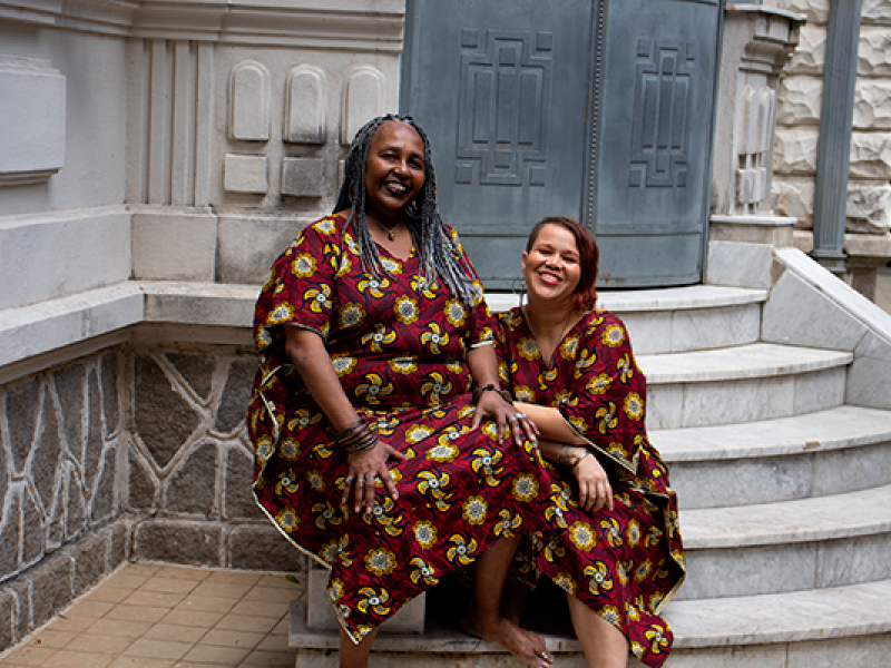 As contadoras de histórias Claudia Marques e Pâmela Cristina sorriem sentadas nos degraus de uma escadaria de pedra diante de um edifício histórico. As duas vestem roupas com a mesma estampa em tons de vinho, amarelo e azul, criando uma identidade visual marcante. O ambiente de arquitetura clássica reforça o clima acolhedor da fotografia.