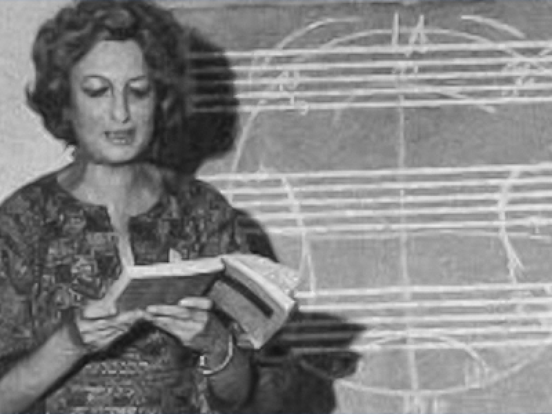 Fotografia em preto e branco da compositora e professora Esther Scliar. Ela aparece lendo um livro aberto, diante de um quadro com diversas linhas e formas geométricas desenhadas a giz. A sombra de seu perfil é projetada sobre o quadro, ao lado das anotações.