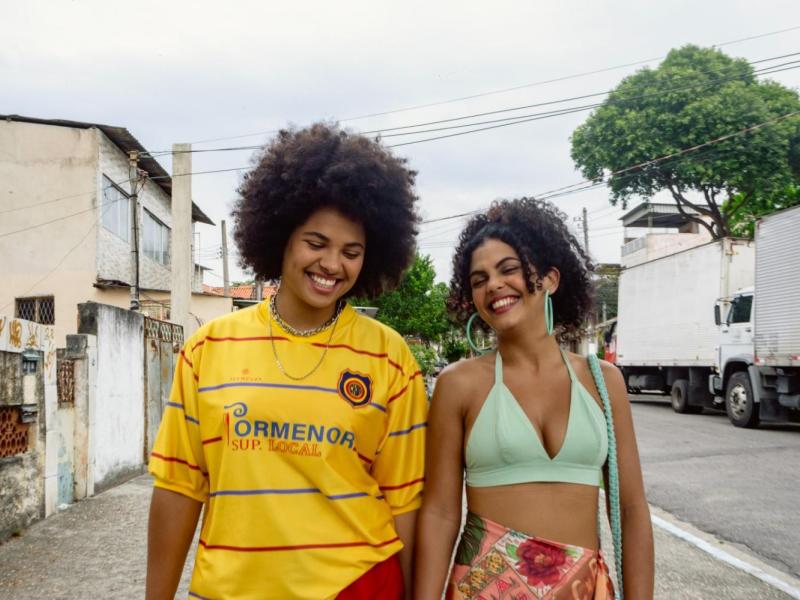 Still de Ficção Suburbana. Duas jovens caminham lado a lado por uma rua de bairro, sorrindo. Uma delas veste camisa amarela de futebol e a outra usa top verde-claro e saia florida. Ao fundo, aparecem casas, muros, árvores, postes, fiação e caminhões estacionados ao longo da rua, sob céu nublado.
