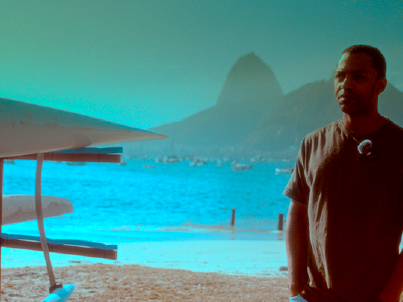 Still do filme Racismo. Um homem aparece em primeiro plano, à direita, usando camiseta escura e olhando para fora do quadro. Ao fundo, vê-se a Baía de Guanabara, com o Pão de Açúcar em destaque entre os morros. À esquerda, parte de uma estrutura com equipamentos náuticos ocupa a lateral da imagem. A cena tem predominância de tons azulados e esverdeados.