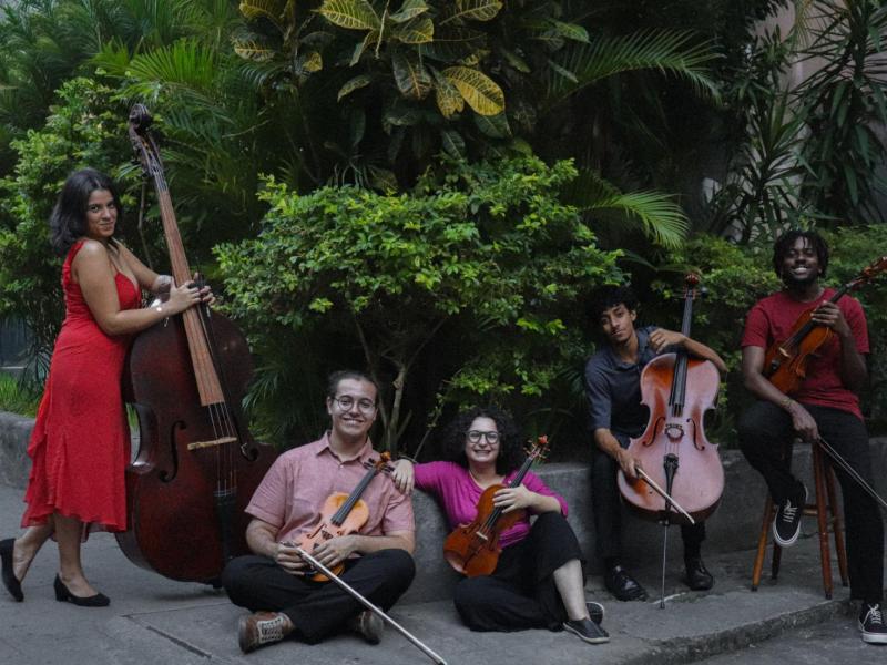 Integrantes do Coletivo Ipê posam com seus instrumentos em uma área externa cercada por vegetação. Os cinco músicos aparecem com instrumentos de cordas e arco de diferentes tamanhos, entre eles contrabaixo e violoncelo. Alguns estão sentados no chão e outros em pé ou sobre um banco, tendo árvores e plantas ao fundo.
