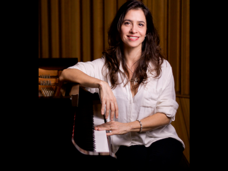 A pianista Marina Spoladore sorri para a câmera enquanto está sentada ao lado de um piano de cauda. Veste camisa branca de mangas dobradas e apoia um dos braços sobre o instrumento, mantendo a outra mão próxima às teclas. Ao fundo, painéis de madeira em tons quentes compõem o ambiente, reforçando a atmosfera acolhedora do retrato.