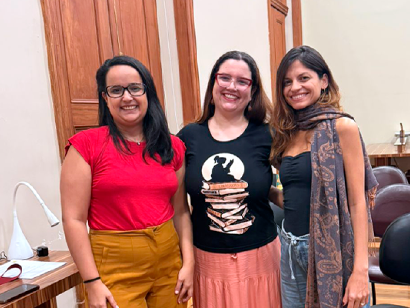 As escritoras e mediadoras Beatriz Ras, Carolina Pessoa e Nicole Ayres posam juntas, sorrindo para a câmera, em uma sala do CCJF. As três aparecem lado a lado, em pé, diante de portas de madeira, vestindo roupas em tons de vermelho, preto, rosa e cinza. Ao fundo, mesas e cadeiras compõem o ambiente.