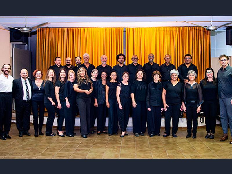Integrantes do Coro Sinfônico da Associação de Canto Coral (ACC) posam reunidos para uma fotografia. O grupo, formado por cerca de vinte e cinco pessoas, está disposto em duas fileiras, com os integrantes da frente no mesmo nível e os de trás sobre uma plataforma. A maioria veste roupas pretas, criando unidade visual, diante de uma grande cortina em tom amarelo-dourado.