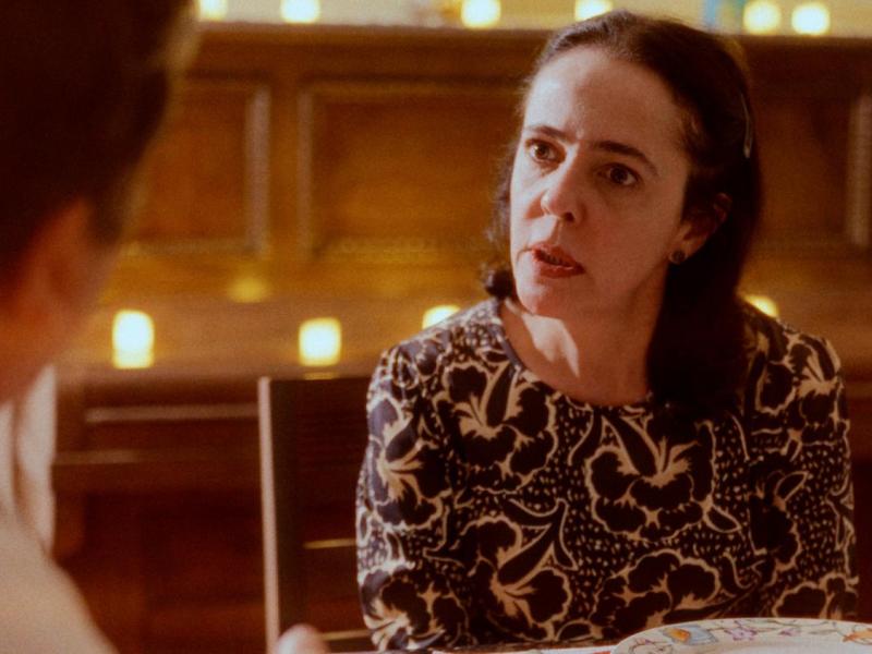 Still do filme O que resta de nós?. A atriz Sílvia Buarque aparece sentada à mesa, voltada para uma pessoa desfocada em primeiro plano. Com expressão atenta e séria, olha diretamente para o interlocutor enquanto parece conversar. Veste uma blusa estampada em preto e branco. Ao fundo, um ambiente iluminado por pequenas luzes em tons quentes reforça a atmosfera intimista da cena.