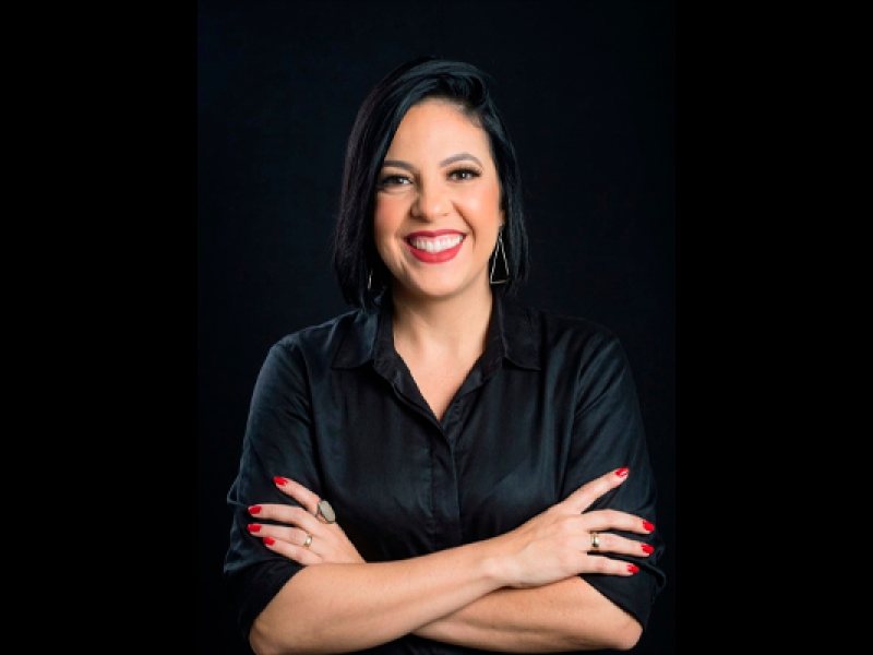 Retrato da professora Tati Vidal. Ela aparece sorrindo para a câmera, com os braços cruzados, cabelos escuros curtos e veste camisa preta. O fundo também é preto, destacando o rosto e as mãos, com unhas pintadas de vermelho.