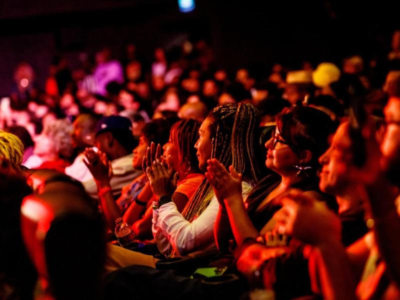 Público durante uma sessão do Encontro de Cinema Negro Zózimo Bulbul. Em primeiro plano, pessoas sentadas lado a lado aplaudem, voltadas para o palco ou a tela fora da imagem. Ao fundo, uma plateia numerosa ocupa o espaço, sob iluminação predominantemente vermelha e alaranjada.