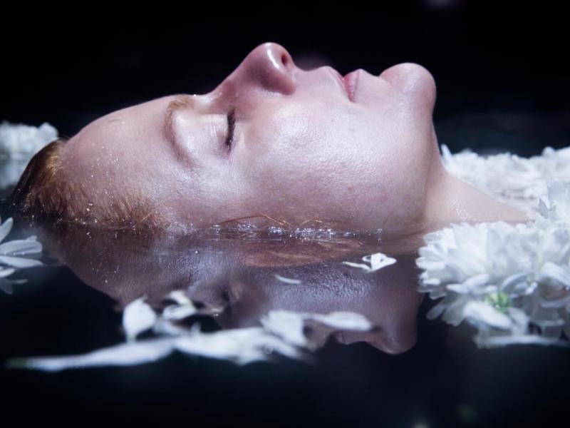Still de curta-metragem da Mostra Vem Ver. Em primeiro plano, uma mulher aparece deitada de olhos fechados, com parte do rosto e dos cabelos mergulhada na água. Flores brancas flutuam ao redor de sua cabeça, e seu rosto se reflete na superfície escura da água.