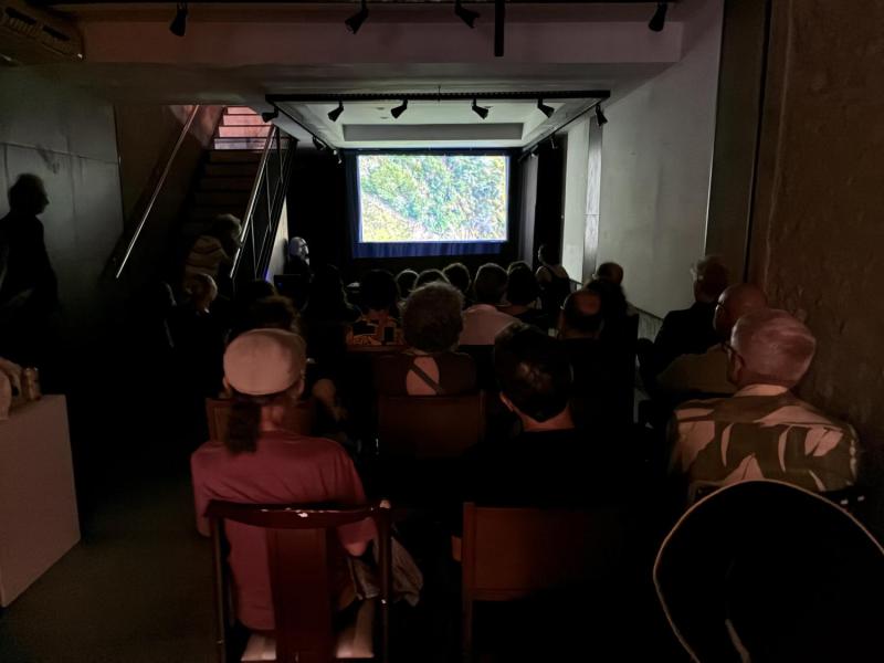Imagem de referência da mostra de videoarte Arca CCJF. Em uma sala escura, o público está sentado em fileiras diante de uma tela na qual é exibido um vídeo. Trilhos com refletores aparecem no teto e, à esquerda, uma escada dá acesso ao piso superior.