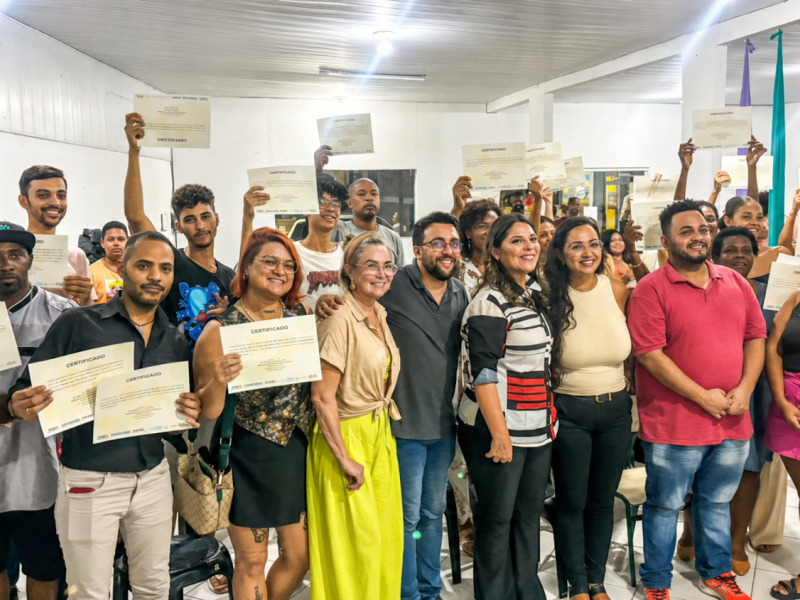 Imagem de referência da Sessão Baixada. Um grande grupo posa reunido em um espaço interno; várias pessoas exibem certificados diante da câmera, enquanto outras aparecem ao centro e ao fundo.