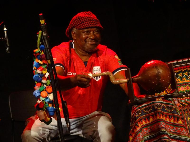 Músico sentado no palco sorri enquanto toca um instrumento de percussão com uma baqueta. Ele veste camisa vermelha e gorro estampado. Ao seu redor, aparecem instrumentos e objetos decorados com tecidos de estampas coloridas, além de um suporte ornamentado com pequenas peças multicoloridas.