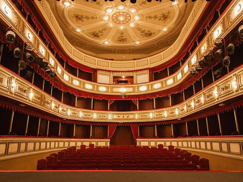 A imagem mostra o interior de um teatro clássico, visto do palco em direção à plateia. O espaço tem assentos estofados em vermelho, três andares de camarotes com detalhes dourados e cortinas vermelhas, além de um teto ornamentado com afrescos e um grande lustre central. A iluminação quente destaca a decoração elegante e simétrica do ambiente.