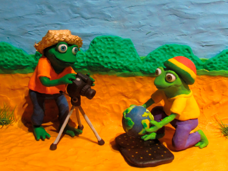 Cena com dois sapos humanoides em estilo de desenho animado, produzidos em massa de modelar. Em um cenário ao ar livre, um deles está diante de uma câmera sobre tripé, enquanto o outro, ajoelhado, segura um pequeno globo terrestre. Ao fundo, aparecem colinas verdes sob um céu azul.
