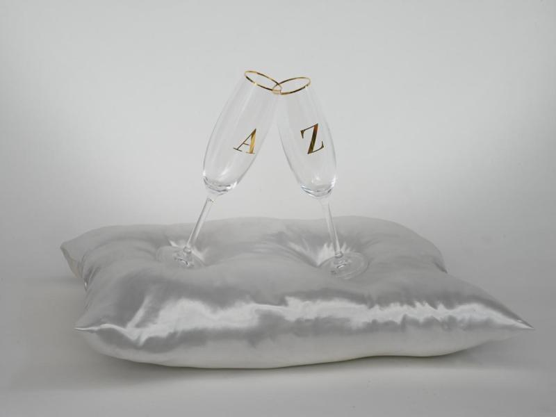 Obra da exposição As Palavras. Duas taças transparentes, com as letras douradas “A” e “Z”, estão inclinadas uma contra a outra, tocando-se pelas bordas. As taças repousam sobre uma almofada branca de tecido acetinado, diante de um fundo claro.