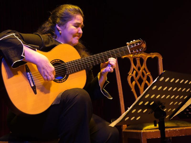 A violonista Mara Lúcia Ribeiro toca sentada no palco, diante de uma estante com partitura. Vestida de preto, ela segura o violão enquanto executa a música, sob iluminação cênica em tons quentes e azulados. Ao lado, há uma cadeira de madeira.