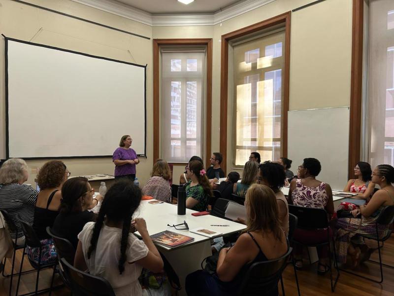Grupo participa de atividade em uma sala, sentado ao redor de mesas dispostas em conjunto. À frente, uma mulher em pé fala aos participantes, que estão voltados em sua direção. O ambiente tem grandes janelas, tela de projeção e quadro branco, e sobre as mesas aparecem livros, cadernos, garrafas e outros materiais.