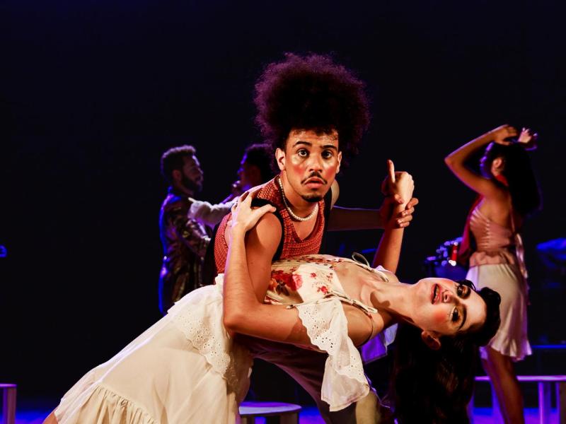 Cena do espetáculo teatral Um Homem Célebre. Em primeiro plano, um ator de cabelos volumosos, usando blusa vermelha sem mangas, sustenta uma atriz em um passo de dança, inclinando-a para trás. Ela veste um figurino claro, com detalhes florais e rendas. O ator olha diretamente para a câmera com expressão de surpresa. Ao fundo, outros artistas aparecem em movimento, sob iluminação cênica em tons de roxo e azul.