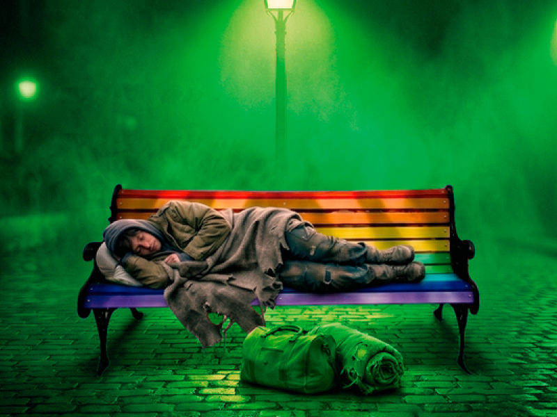 Em uma cena noturna envolta por névoa verde, uma pessoa com roupas desgastadas dorme encolhida sobre um banco pintado com as cores do arco-íris. No chão de pedras, à frente do banco, estão uma mochila e um cobertor enrolado. Ao fundo, postes de iluminação atravessam a névoa.