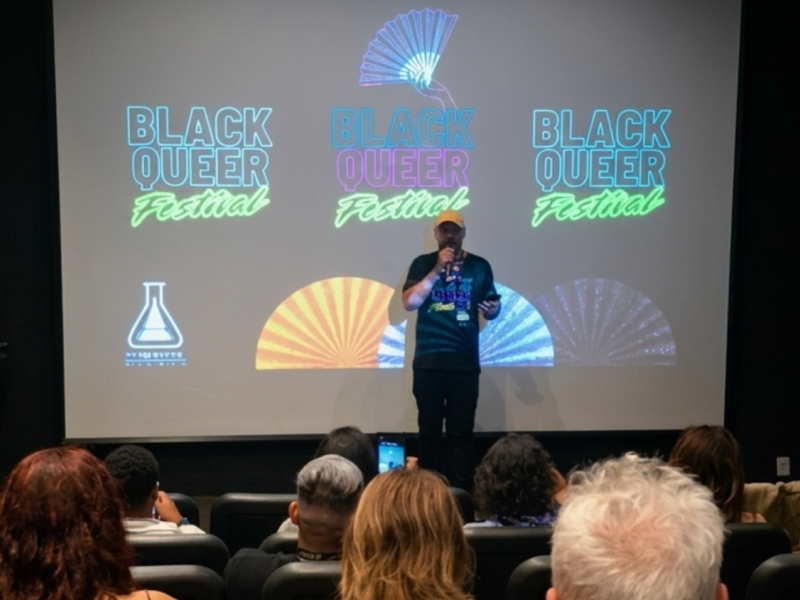 Foto do Black Queer Festival. Em uma sala de cinema, uma pessoa de boné e camiseta preta fala ao microfone diante de uma plateia sentada. Ao fundo, uma grande tela exibe três vezes o logotipo colorido “Black Queer Festival”, acompanhado de ilustrações de leques.