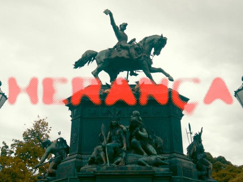 Cartaz do curta-metragem Herança. Fotografia de um monumento equestre, com a estátua de um homem montado a cavalo, erguendo um dos braços. Na base do monumento, aparecem esculturas de figuras humanas sentadas. Sobre a imagem, o título “Herança” está escrito em letras grandes e vermelhas, com efeito pontilhado. Ao fundo, céu nublado e árvores.