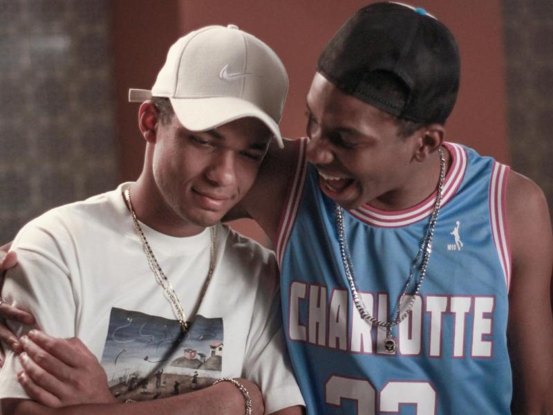 Cena do filme Nosso Sonho. À esquerda, o ator Juan Paiva, no papel de Buchecha, usa boné branco, camiseta clara e corrente dourada. Ao seu lado, Lucas Penteado, que interpreta Claudinho, veste uma regata azul de basquete e boné preto virado para trás. Lucas abraça Juan pelos ombros e sorri enquanto fala com ele, que mantém o olhar abaixado e um sorriso discreto.