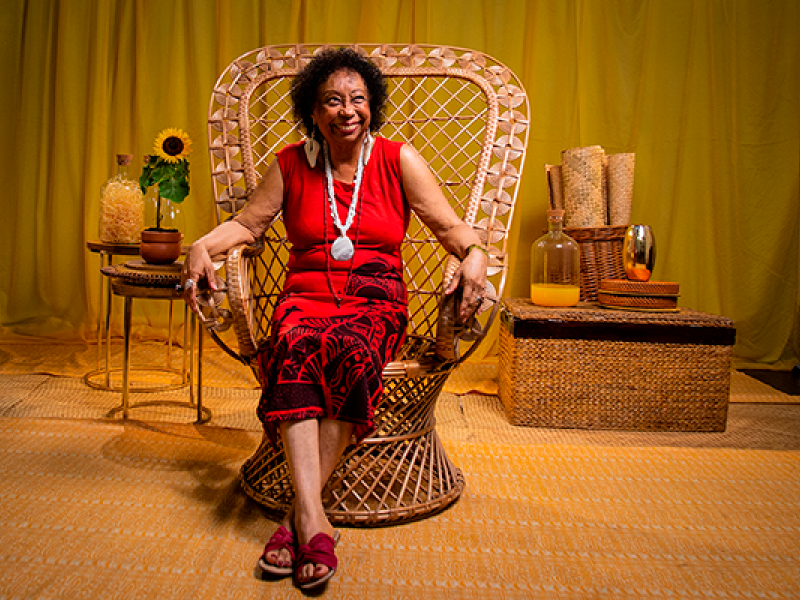 Mulher negra de cabelos curtos e cacheados sorri, sentada em uma grande poltrona de vime trançado. Ela veste roupa vermelha com estampas pretas, usa colares compridos e sandálias vermelhas. O cenário, em tons de amarelo e dourado, é composto por cortinas, tapete e móveis de fibras naturais. Nas laterais, pequenos arranjos decorativos incluem um girassol, vasos, garrafas e cestos.