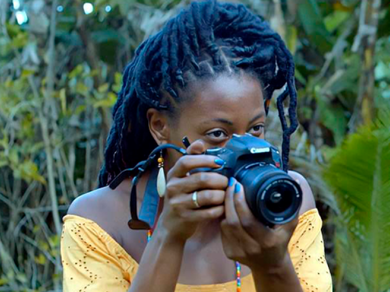 Cena do curta-metragem Como Matar uma Boneca. Em meio à vegetação, uma jovem negra de cabelos longos em tranças segura uma câmera fotográfica Canon diante do rosto, preparando-se para fotografar. Ela veste uma blusa amarela de ombros descobertos e usa brincos compridos.
