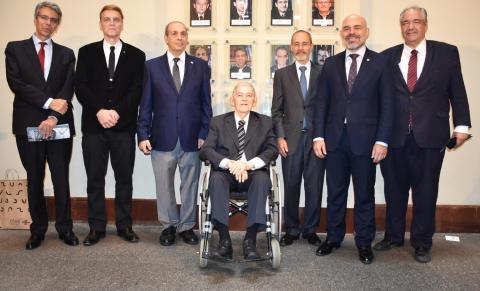 Oito homens posam lado a lado em frente a uma parede com a inscrição “Galeria dos Diretores do CCJF” e retratos emoldurados. Todos usam ternos formais. Um deles está sentado em uma cadeira de rodas no centro do grupo. Os demais estão em pé, sorrindo. A cena ocorre em ambiente interno, com iluminação clara e piso de pedra cinza.