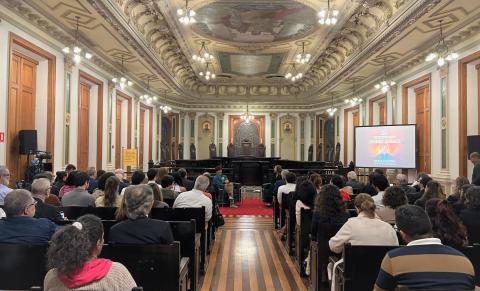 Auditório nobre com teto ornamentado e afrescos, paredes com molduras e luminárias pendentes. O espaço está cheio de pessoas sentadas, voltadas para o palco ao fundo, onde ocorre uma palestra. À direita, há um telão com a imagem do jornalista Guga Chacra e os dizeres “Encontros Acadêmicos”. O ambiente é solene e bem iluminado.