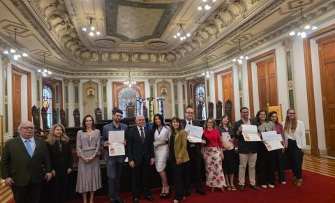 Foto em um grande salão histórico, com teto alto e ornamentado, lustres acesos, vitrais e cadeiras de madeira ao fundo. Um grupo de cerca de 14 pessoas, mulheres e homens adultos, está alinhado sobre um tapete vermelho, alguns segurando certificados. Eles posam sorrindo para a câmera, em clima de cerimônia ou premiação oficial.