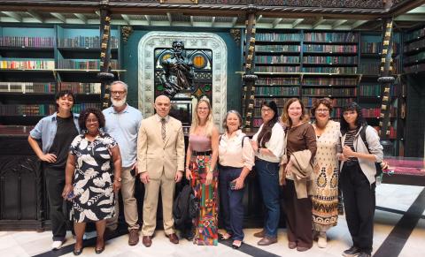 Grupo de dez pessoas posa lado a lado em uma grande biblioteca histórica. Ao fundo, estantes altas e circulares repletas de livros sobem até o teto, com estrutura ornamentada de ferro e madeira e um grande lustre central. O grupo sorri para a câmera. Há diversidade de idades, gêneros e tons de pele. Vestem roupas casuais e sociais. O piso é metálico vazado e a iluminação é quente, destacando o ambiente elegante e cultural.