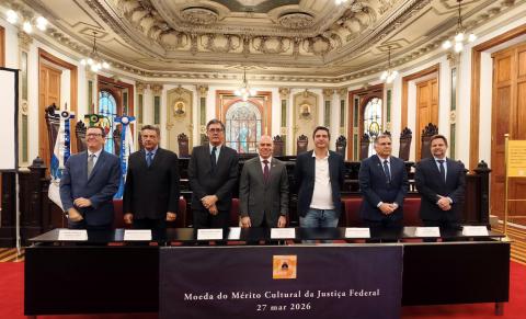 Foto de cerimônia oficial em um salão histórico ricamente decorado, com teto ornamentado, lustres e vitrais ao fundo. Sete homens adultos estão em pé, alinhados atrás de uma mesa longa, usando trajes formais (ternos e gravatas, um em roupa mais casual). À frente da mesa, uma placa preta com o texto: “Moeda do Mérito Cultural da Justiça Federal – 27 mar 2026”. O ambiente é solene, com bandeiras laterais e cadeiras de madeira ao fundo.
