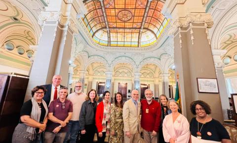 Fotografia de grupo em um amplo salão histórico de arquitetura ornamentada. Doze pessoas posam lado a lado, sorrindo para a câmera, em frente a colunas altas e arcos decorados em tons claros. Ao fundo, destaca-se um grande vitral colorido no teto, que ilumina o ambiente com luz suave. Os participantes usam roupas variadas, em tons neutros e vermelhos. À direita, vê-se parcialmente uma bandeira do Brasil. A imagem transmite um clima institucional e de confraternização.