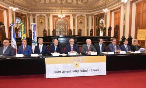 Fotografia da Sala de Sessões do Centro Cultural Justiça Federal. Em primeiro plano, nove autoridades, entre homens e uma mulher, estão sentados lado a lado atrás de uma longa mesa preta, durante uma cerimônia institucional. À frente da mesa, uma faixa exibe a inscrição “Centro Cultural Justiça Federal – 25 anos”. Ao fundo, destacam-se os vitrais históricos, cadeiras de madeira, bandeiras e a arquitetura ornamentada do salão, com teto decorado, luminárias suspensas e detalhes dourado, verde e creme.