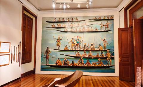 Sala expositiva com piso de madeira polida e paredes claras. Ao centro da parede principal há um grande painel pintado em tons de azul, retratando diversos indígenas em longas canoas sobre a água, alguns remando, sentados ou em pé com arcos e lanças. No primeiro plano, vê-se uma canoa de madeira real. À esquerda, remos e objetos indígenas estão expostos na parede. Portas e janelas de madeira escura emolduram o ambiente, iluminado por refletores no teto.