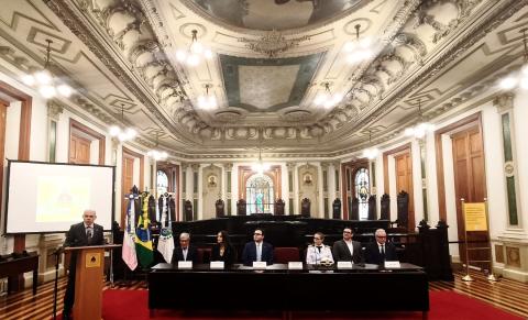 Foto de cerimônia na Sala de Sessões do CCJF, com teto ricamente ornamentado e lustres. Ao fundo, vitrais e cadeiras de madeira. À frente, uma mesa reúne sete pessoas sentadas, entre elas uma mulher uniformizada, e há bandeiras ao lado. À esquerda, um homem de terno fala ao microfone, diante de um telão. O piso e o tapete são vermelhos.