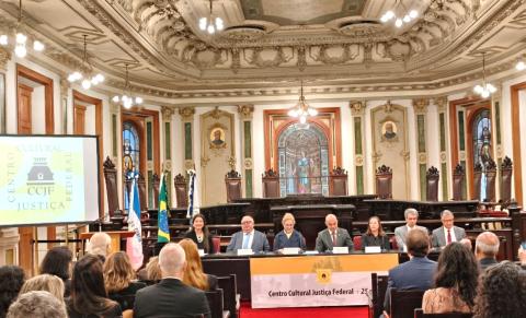 Em um salão histórico e ornamentado, seis pessoas estão sentadas à mesa de um evento, diante de uma plateia. Ao fundo, há vitrais, esculturas, pinturas, bandeiras do Brasil e outras, além de detalhes dourados no teto e nas paredes. Um telão à esquerda exibe o logotipo do Centro Cultural Justiça Federal.