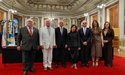 Fotografia de sete pessoas, quatro homens e três mulheres, posando lado a lado em um salão solene, sobre um tapete vermelho. Ao fundo, há cadeiras de madeira escura, vitrais coloridos, bandeiras e paredes com detalhes ornamentais dourados. O teto é ricamente decorado com relevos e lustres. Os participantes vestem trajes formais e sorriem para a foto