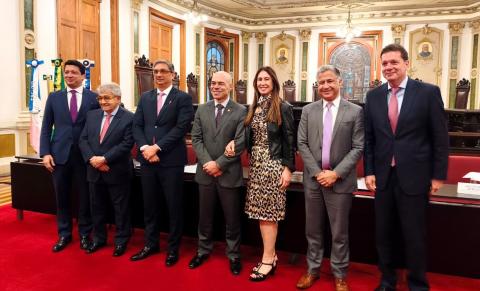 Seis pessoas posam lado a lado em um salão suntuoso, com piso vermelho, paredes ornamentadas e teto ricamente decorado. Há quatro homens de terno à esquerda e à direita, outro homem ao centro e uma mulher entre eles, usando vestido estampado de oncinha e jaqueta preta. Ao fundo, veem-se portas de madeira, vitrais, cadeiras e bandeiras.