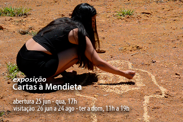 Mulher de cabelos longos e escuros, com roupa preta, agachada em um terreno seco e avermelhado, desenha no chão com o que parece ser pó ou grãos claros. A figura traçada lembra uma cruz estilizada ou um símbolo antropomórfico. A cena sugere um gesto ritualístico ou artístico, em forte contraste com o solo árido ao redor, evocando temas de memória, ancestralidade ou intervenção poética no espaço.Título: exposição Cartas à Mendieta. abertura 25 jun - qua, 17h | visitação  26 jun a 24 ago - ter a dom, 11 a 19h